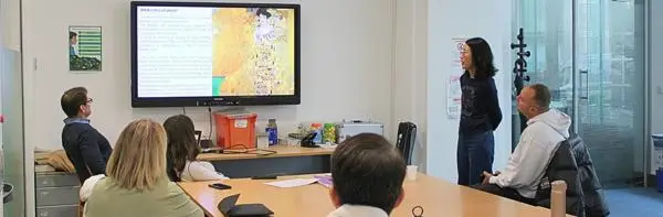 Small group seated around a table watching a presentation on a screen about an artwork, while a presenter stands beside the display in a classroom or workshop setting