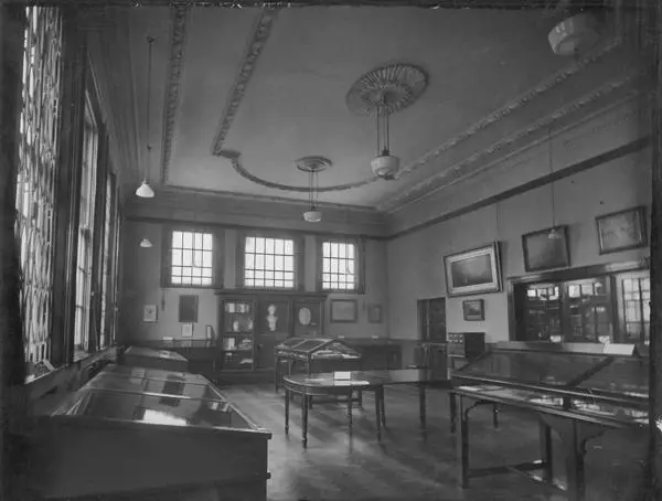 A room with display cabinets for objects, pictures on the walls, and three high windows at the far end.