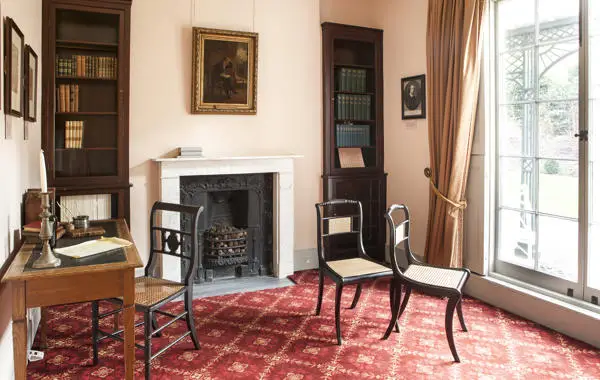 A photograph of a Regency interior with a writing desk and chairs and framed pictures on the walls.